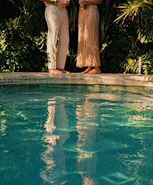 legs of two barefoot people stading above a reflecting blue-water pool with tropical greenery