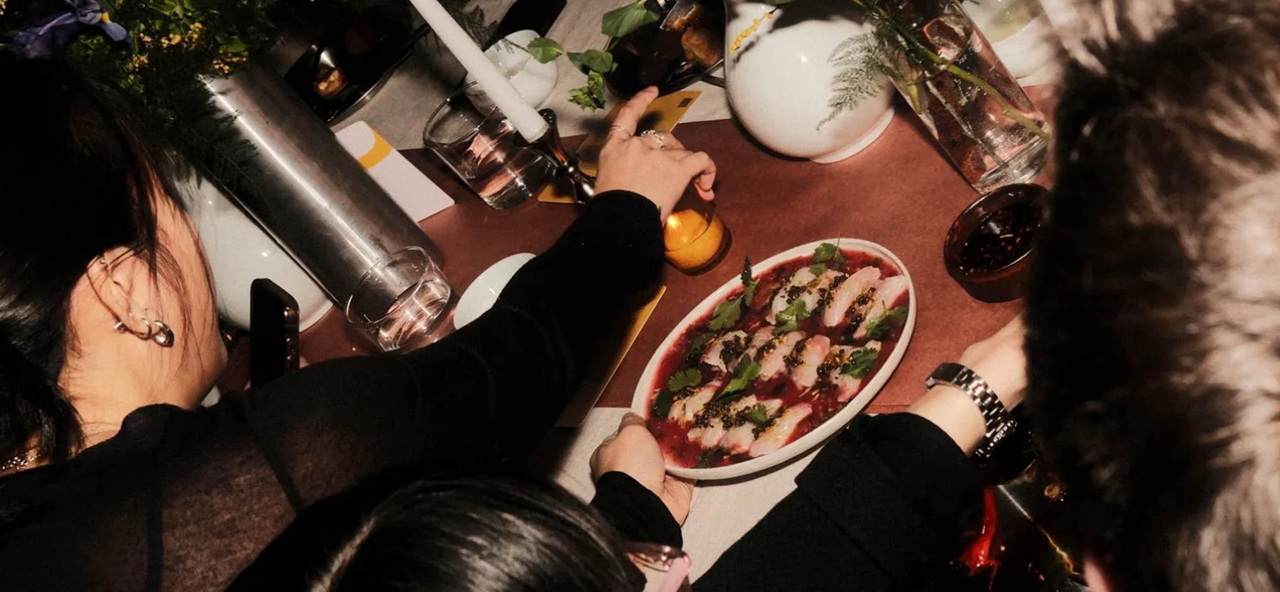 woman reaching for a lit candle while being served food at a large dining table