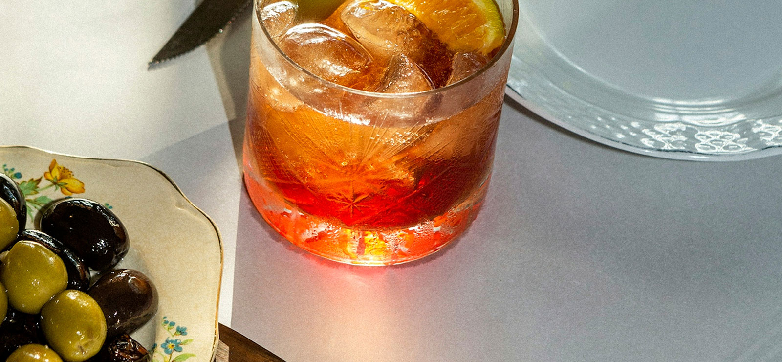 bright orange cocktail with ice in a glass, next to a plate of green and black olives
